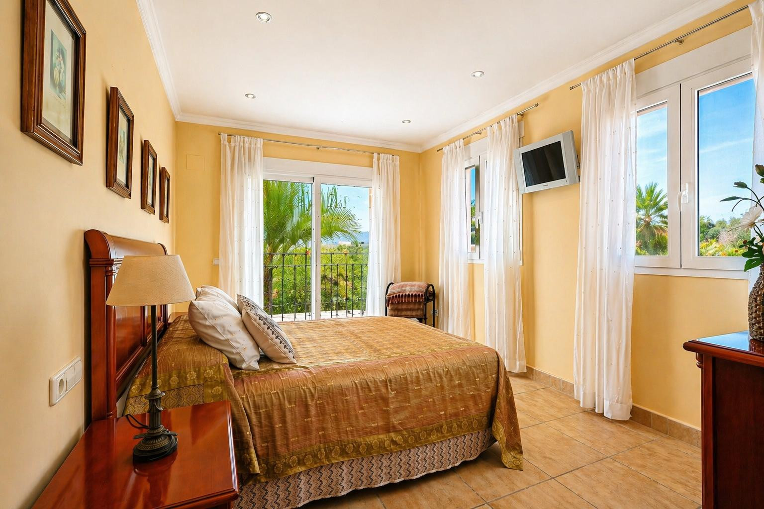 Chambre lumineuse avec lit double et tête de lit en bois, sol carrelé et accès direct à un balcon privé avec vue sur les palmiers et la verdure.