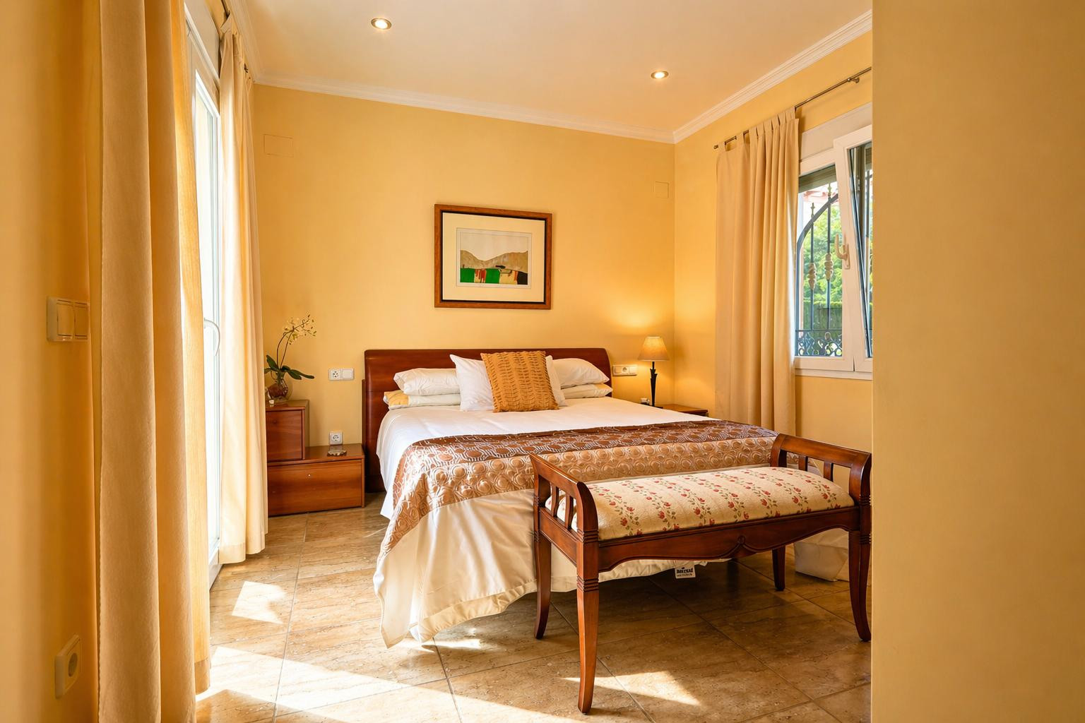Chambre lumineuse avec lit double, tête de lit en bois et sol carrelé. Elle offre un accès direct à l'extérieur et une fenêtre à barreaux décoratifs.