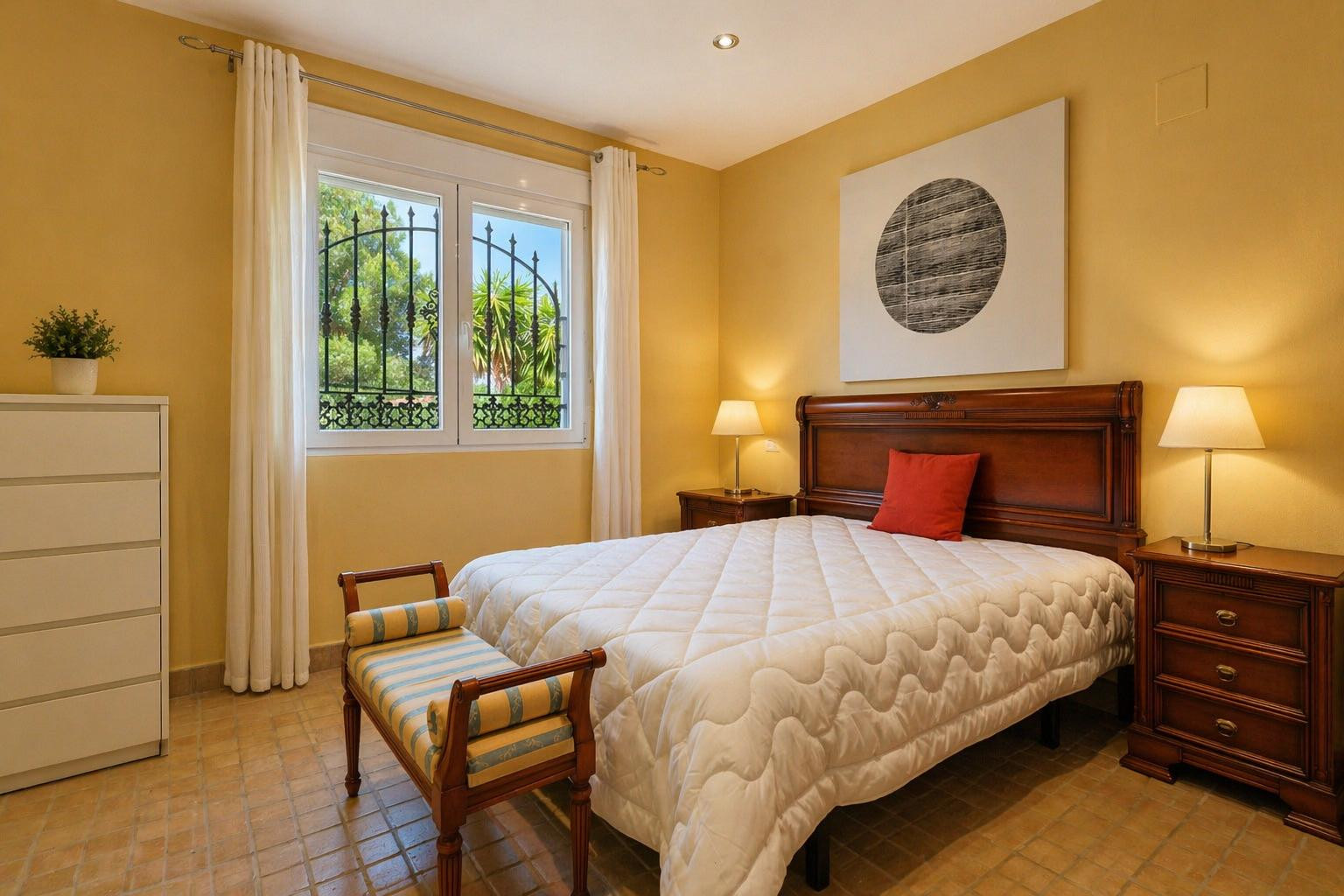 Chambre lumineuse avec lit en bois massif, tables de chevet assorties et carrelage en terre cuite. La fenêtre à barreaux en fer forgé donne sur le jardin.