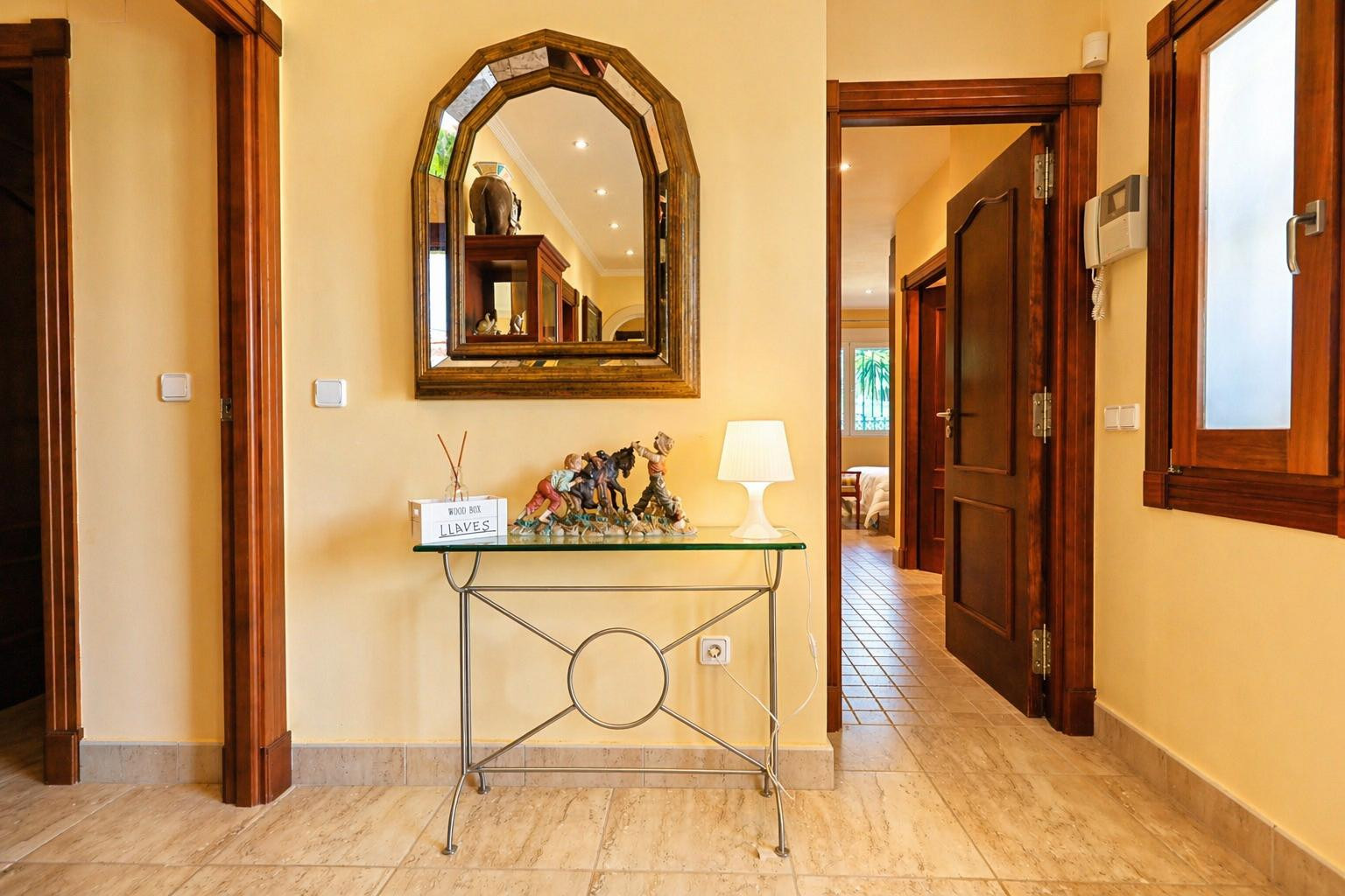 Couloir d'entrée lumineux avec murs jaune clair et sol carrelé. Il présente un miroir arqué en bois au-dessus d'une console en verre, des portes en bois foncé et un interphone.