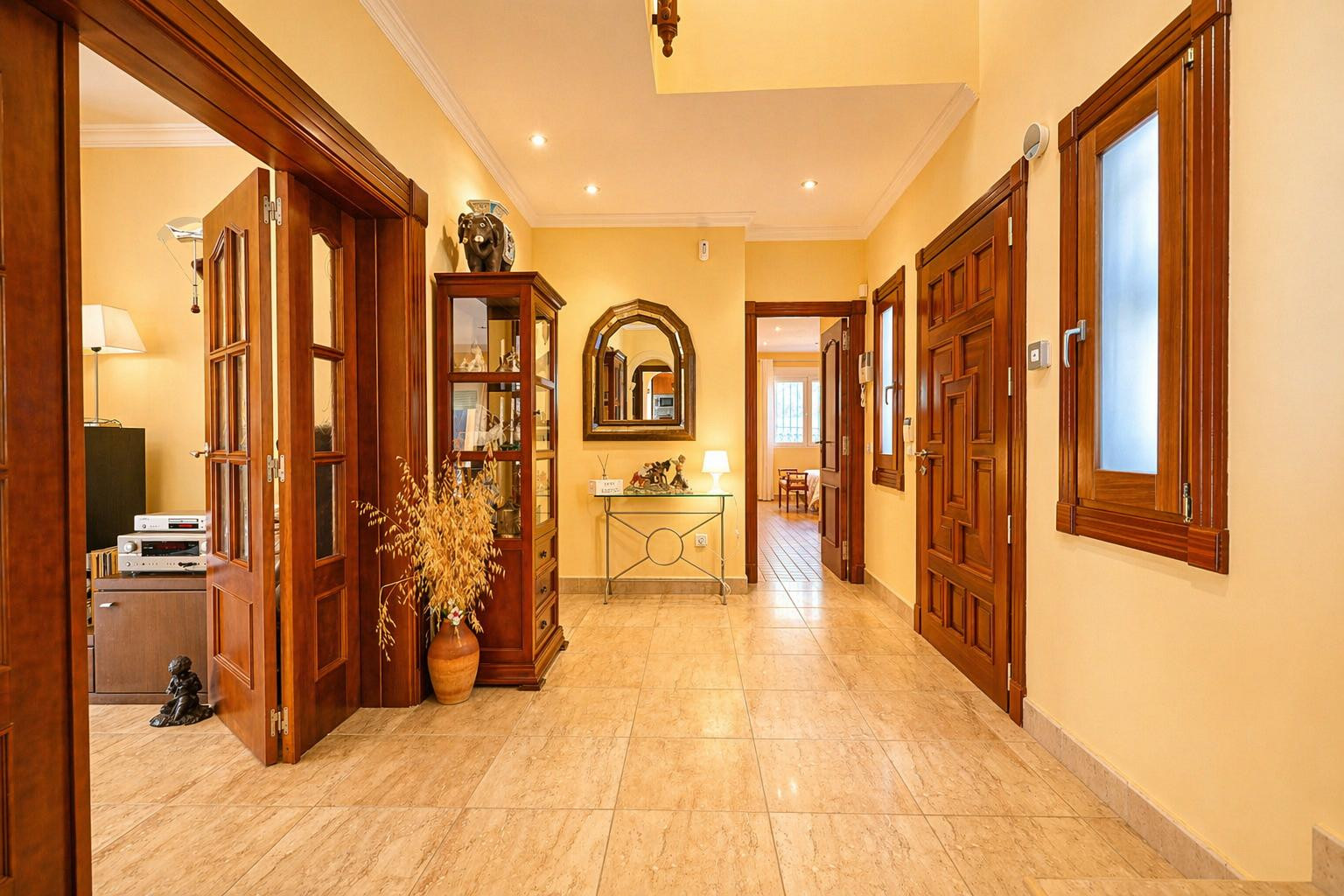 Couloir lumineux aux murs jaune chaud et carrelage clair. Il présente des portes en bois foncé, une vitrine décorative et une console avec un miroir arqué.