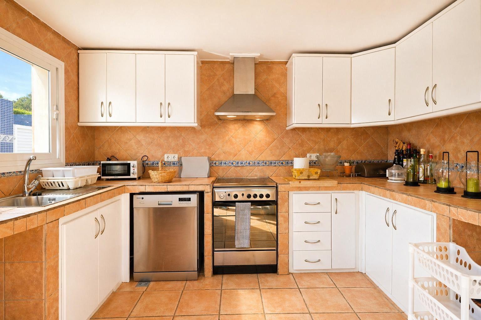 Cuisine entièrement équipée avec carrelage de sol et crédence en terre cuite, meubles hauts et bas blancs, et appareils électroménagers en acier inoxydable. Lumière naturelle par la fenêtre.