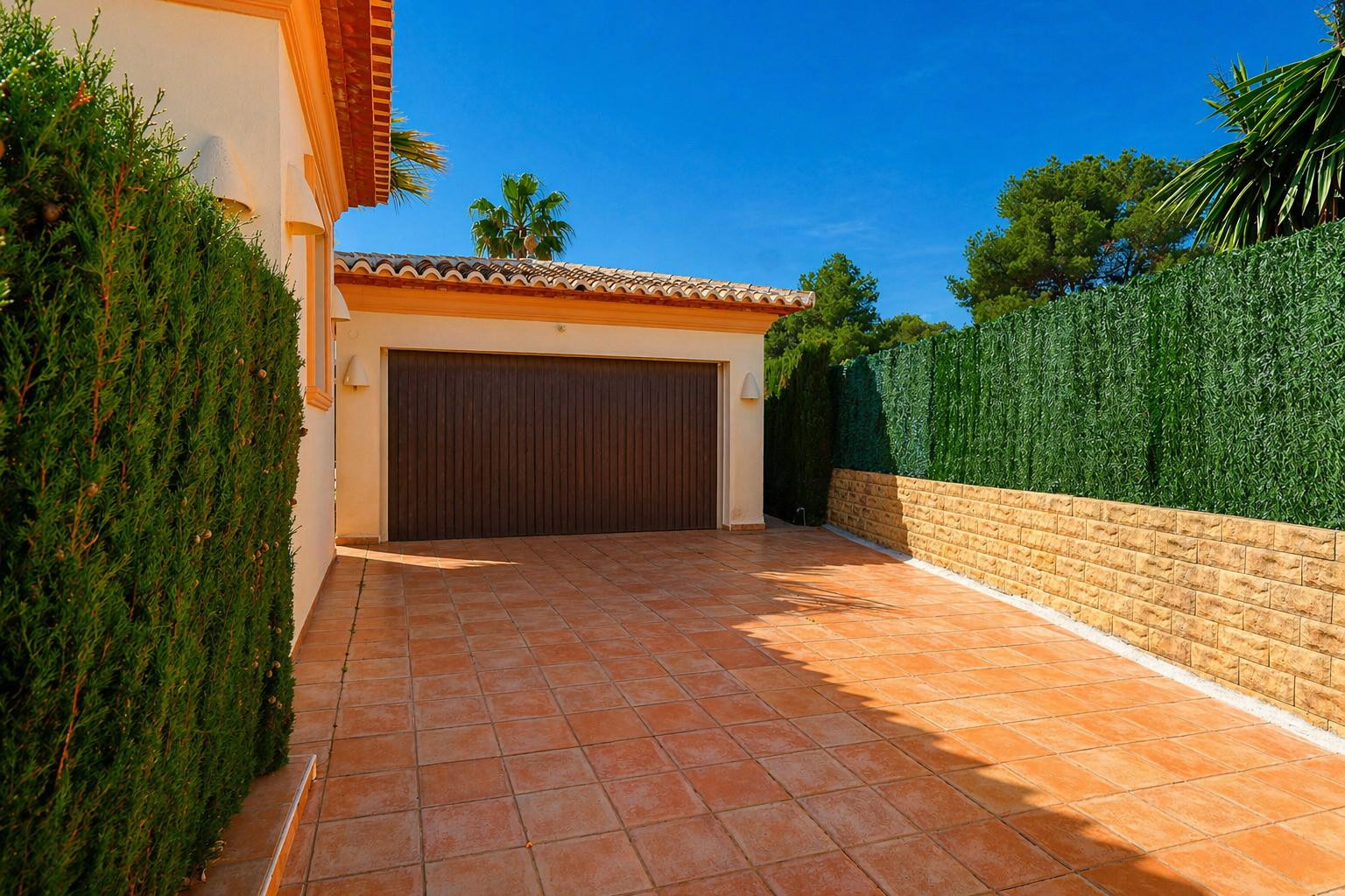 Garage privé avec porte en bois foncé et allée carrelée en terre cuite, bordé d'une haie naturelle et d'un mur en pierre avec écran vert artificiel, sous un ciel bleu clair.