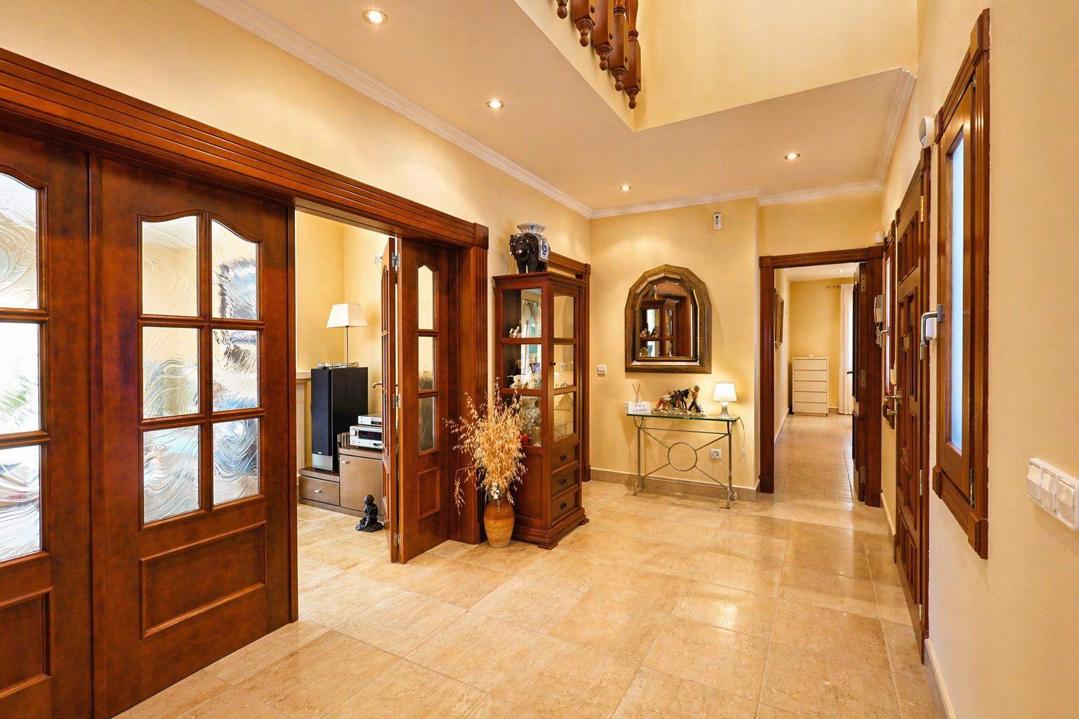 Le couloir lumineux présente un sol carrelé clair et des spots encastrés. Des portes en bois foncé avec vitres décorées mènent aux pièces adjacentes, avec une console et vitrine.