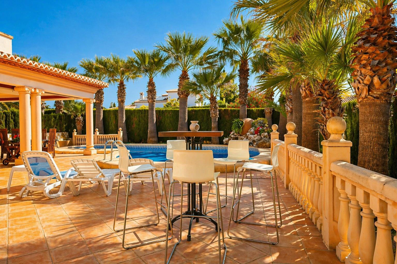 Le patio extérieur privé offre une table haute avec tabourets, des chaises longues et une balustrade sur carreaux de terre cuite. La piscine est bordée de palmiers matures et d'une haie luxuriante.