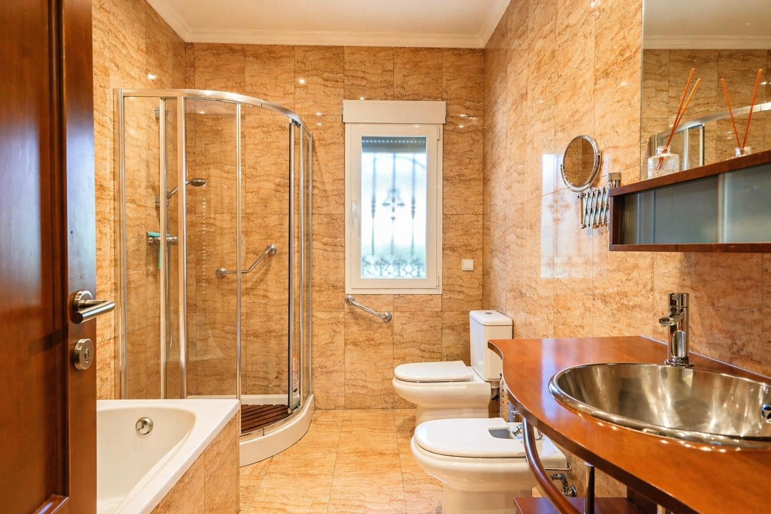 Salle de bain complète avec carrelage terre cuite, baignoire, douche en verre courbé avec barres d'appui, WC, bidet et meuble vasque en bois avec lavabo inox.