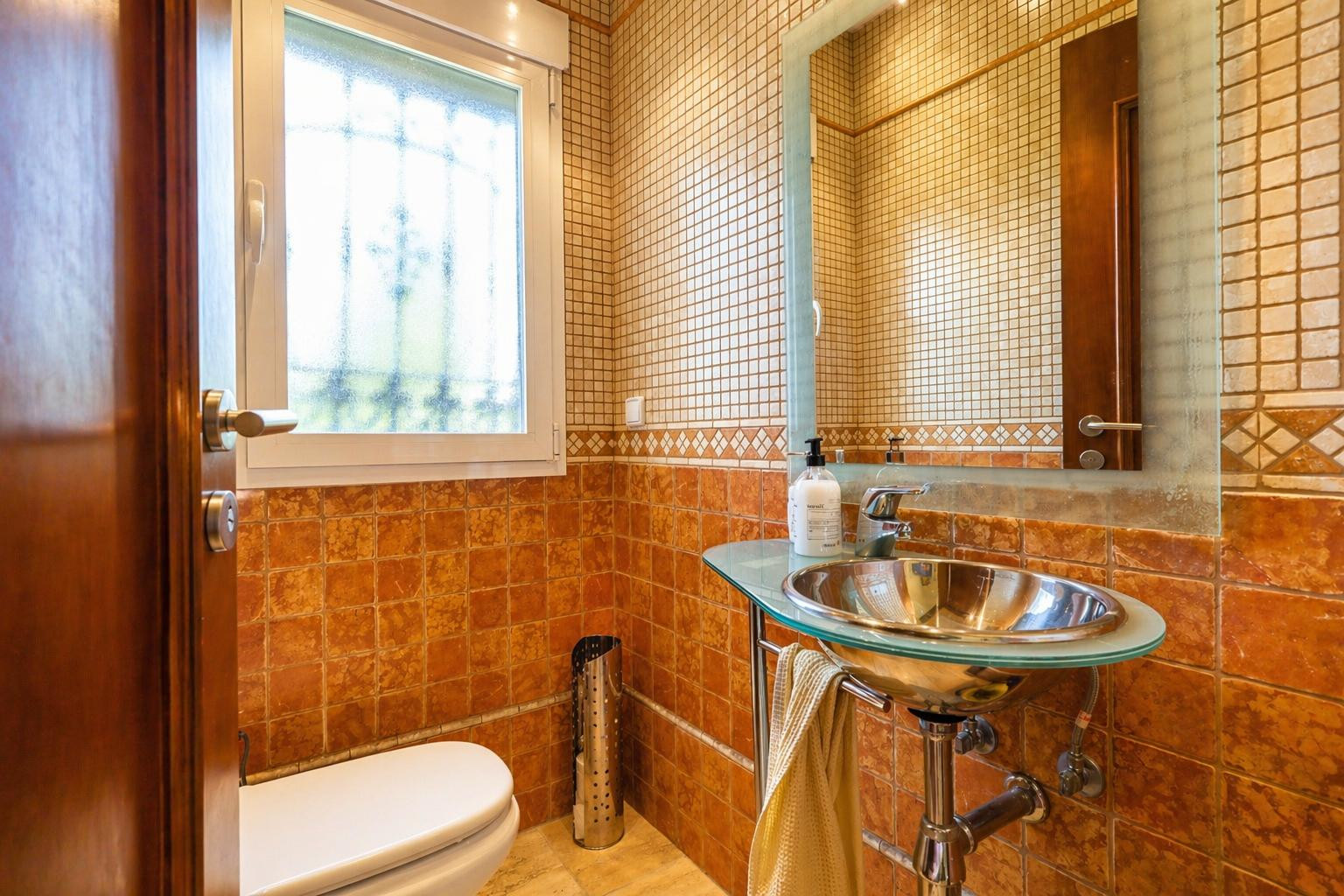 Toilettes d'invités avec carrelage terre cuite et mosaïque, lavabo design à vasque métallique et plateau en verre, et fenêtre lumineuse.