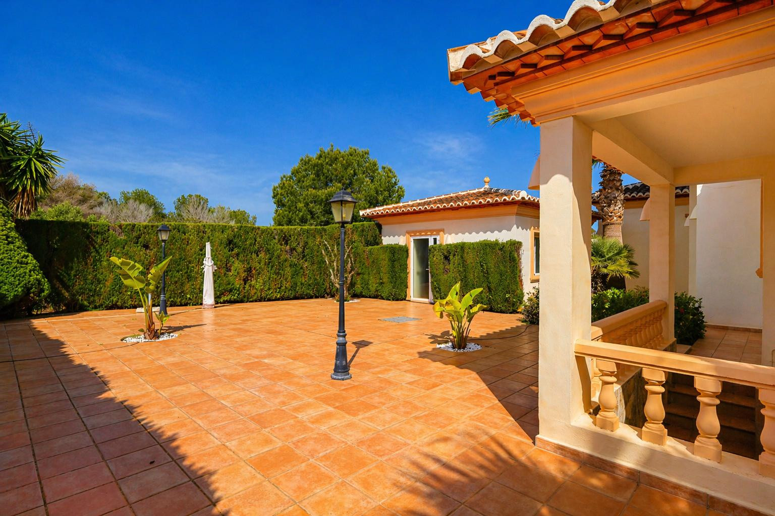 Vaste patio extérieur carrelé en terre cuite, délimité par une haie verte dense, avec des lampadaires classiques et un porche couvert à balustrade et toit tuilé.