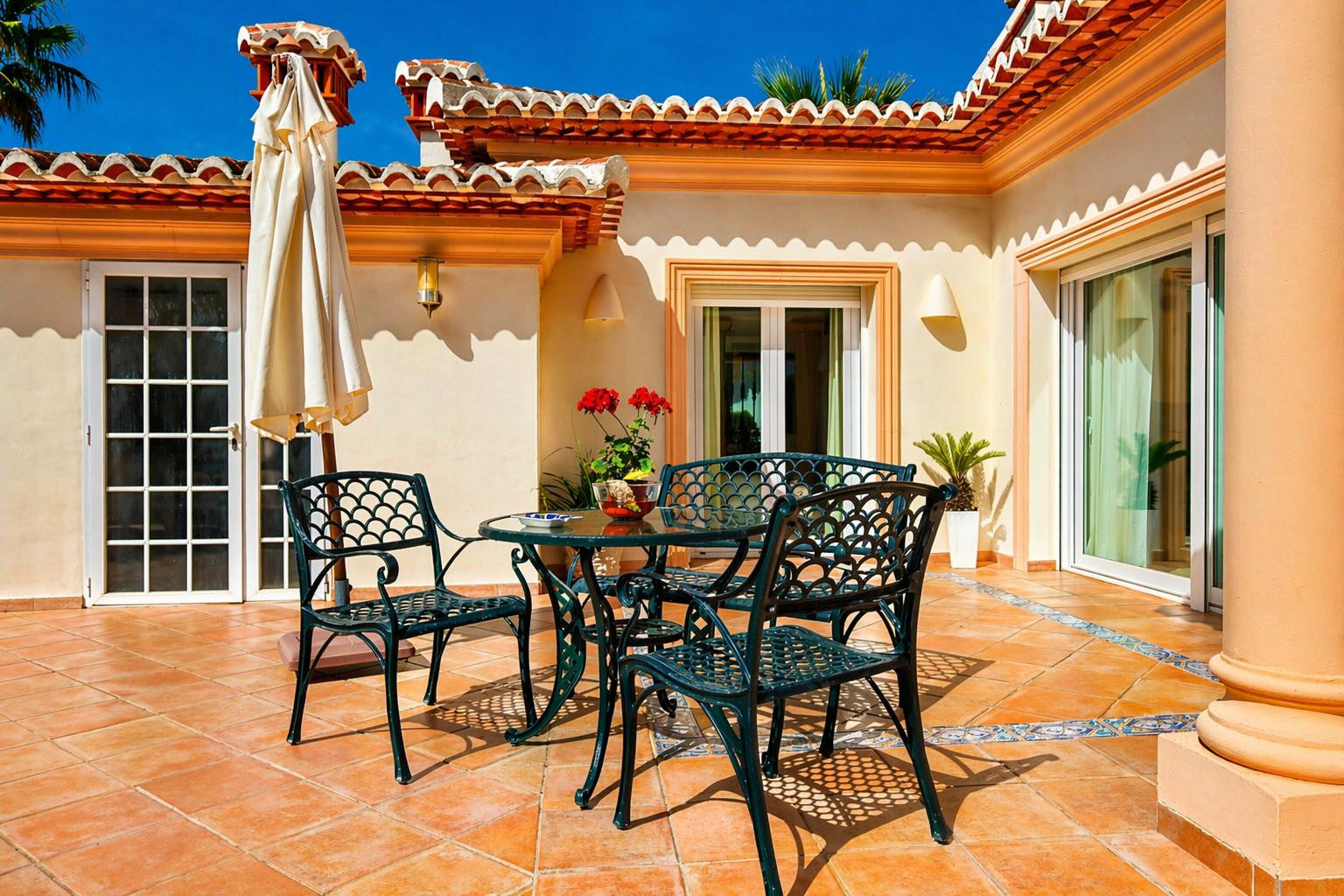 Vaste terrasse extérieure avec sol en terre cuite et bordure en carreaux décoratifs. Elle présente un ensemble de salle à manger en métal vert foncé et une architecture méditerranéenne.