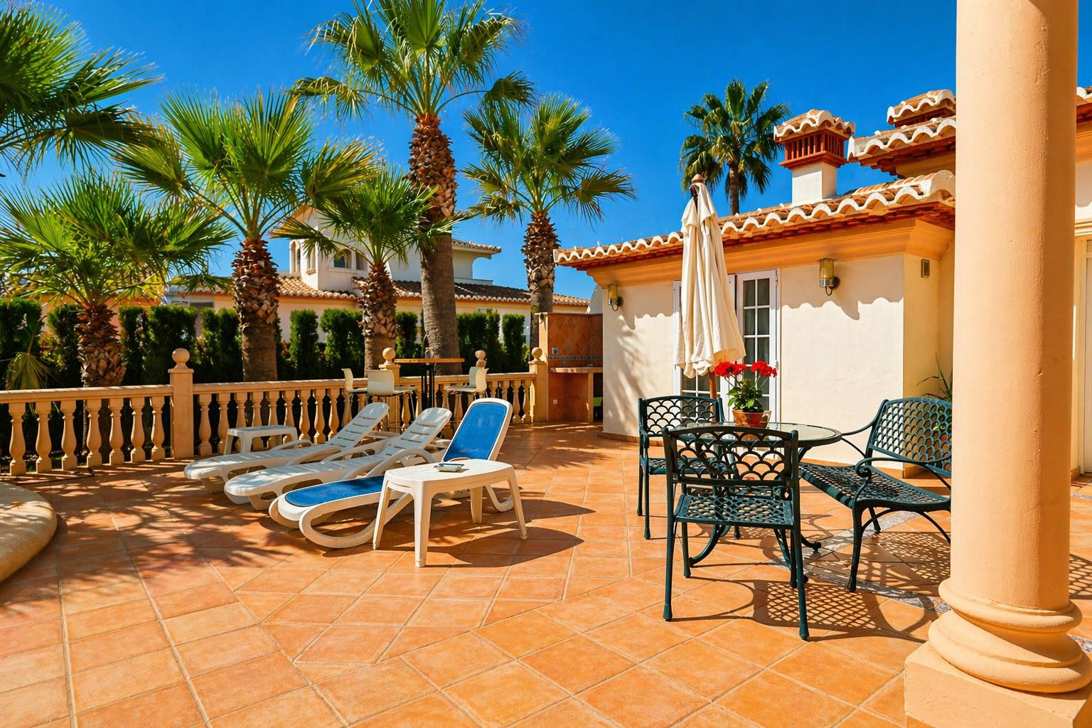 Vaste terrasse extérieure carrelée de terre cuite, aménagée avec des chaises longues et un ensemble repas en métal sous un parasol. Des palmiers et une balustrade en pierre bordent l'espace.