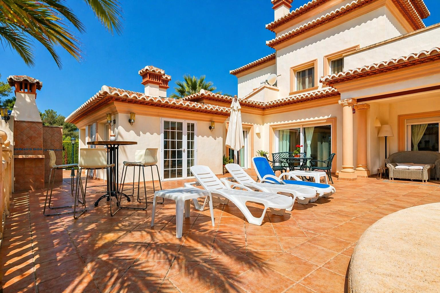 Vaste terrasse extérieure en terre cuite, équipée de chaises longues et d'un espace barbecue intégré, avec accès direct à la villa.