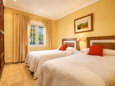 Chambre double avec lits jumeaux, têtes de lit en bois et coussins orange. Elle dispose d'une armoire intégrée, d'un sol carrelé et d'une fenêtre à barreaux.