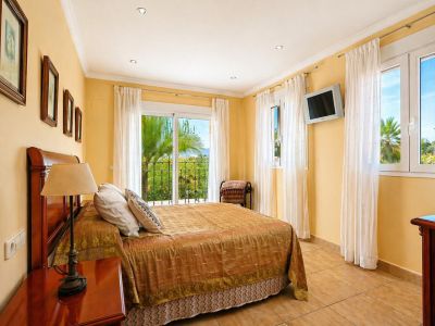 Chambre lumineuse avec lit double et tête de lit en bois, sol carrelé et accès direct à un balcon privé avec vue sur les palmiers et la verdure.