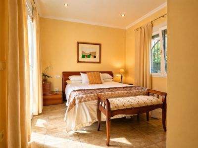 Chambre lumineuse avec lit double, tête de lit en bois et sol carrelé. Elle offre un accès direct à l'extérieur et une fenêtre à barreaux décoratifs.