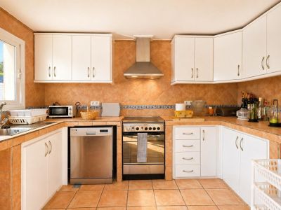 Cuisine entièrement équipée avec carrelage de sol et crédence en terre cuite, meubles hauts et bas blancs, et appareils électroménagers en acier inoxydable. Lumière naturelle par la fenêtre.