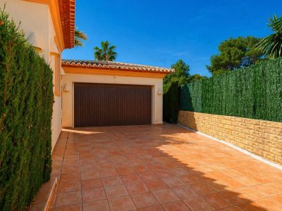 Garage privé avec porte en bois foncé et allée carrelée en terre cuite, bordé d'une haie naturelle et d'un mur en pierre avec écran vert artificiel, sous un ciel bleu clair.