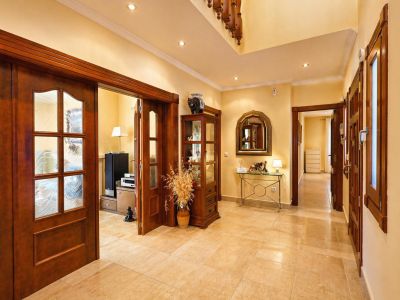 Le couloir lumineux présente un sol carrelé clair et des spots encastrés. Des portes en bois foncé avec vitres décorées mènent aux pièces adjacentes, avec une console et vitrine.