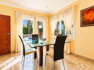 Salle à manger lumineuse avec table en verre, chaises modernes et carrelage au sol à motif géométrique. De grandes fenêtres donnent sur le jardin privé et la piscine.