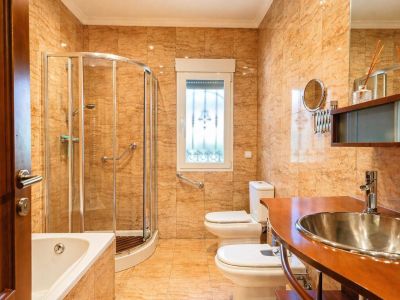 Salle de bain complète avec carrelage terre cuite, baignoire, douche en verre courbé avec barres d'appui, WC, bidet et meuble vasque en bois avec lavabo inox.