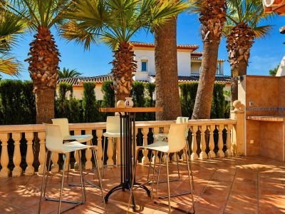 Terrasse extérieure avec carrelage en terre cuite, balustrade classique et un ensemble table haute avec tabourets. Un espace barbecue intégré et des palmiers procurent de l'ombre.