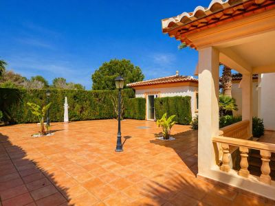 Vaste patio extérieur carrelé en terre cuite, délimité par une haie verte dense, avec des lampadaires classiques et un porche couvert à balustrade et toit tuilé.