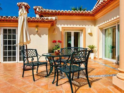 Vaste terrasse extérieure avec sol en terre cuite et bordure en carreaux décoratifs. Elle présente un ensemble de salle à manger en métal vert foncé et une architecture méditerranéenne.