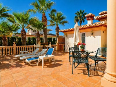 Vaste terrasse extérieure carrelée de terre cuite, aménagée avec des chaises longues et un ensemble repas en métal sous un parasol. Des palmiers et une balustrade en pierre bordent l'espace.