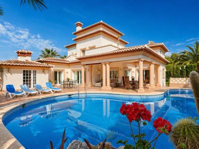 Vue extérieure de la villa avec piscine incurvée en mosaïque bleue et transats. La terrasse couverte à colonnes propose des espaces salon et salle à manger extérieurs, avec accès direct au jardin.