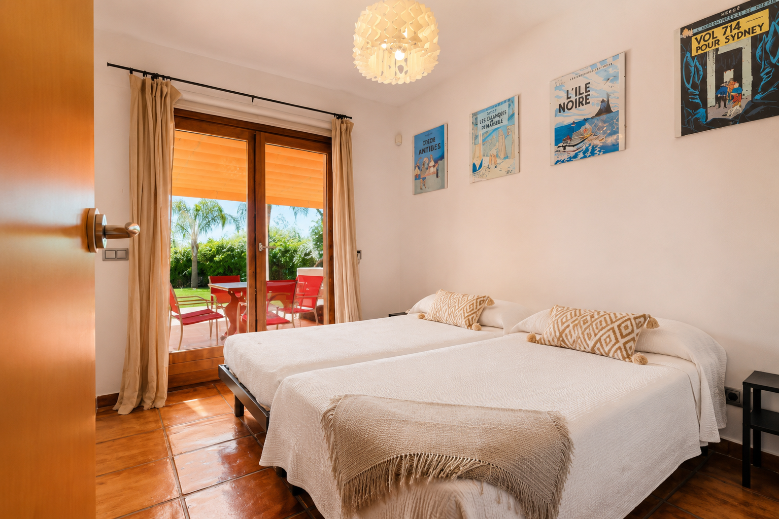 Chambre double avec carrelage en terre cuite et accès direct à une terrasse extérieure. La porte vitrée mène à un jardin verdoyant avec mobilier rouge et palmier. Chambre double avec carrelage en terre cuite et accès direct à une terrasse extérieure. La porte vitrée mène à un jardin verdoyant avec mobilier rouge et palmier.