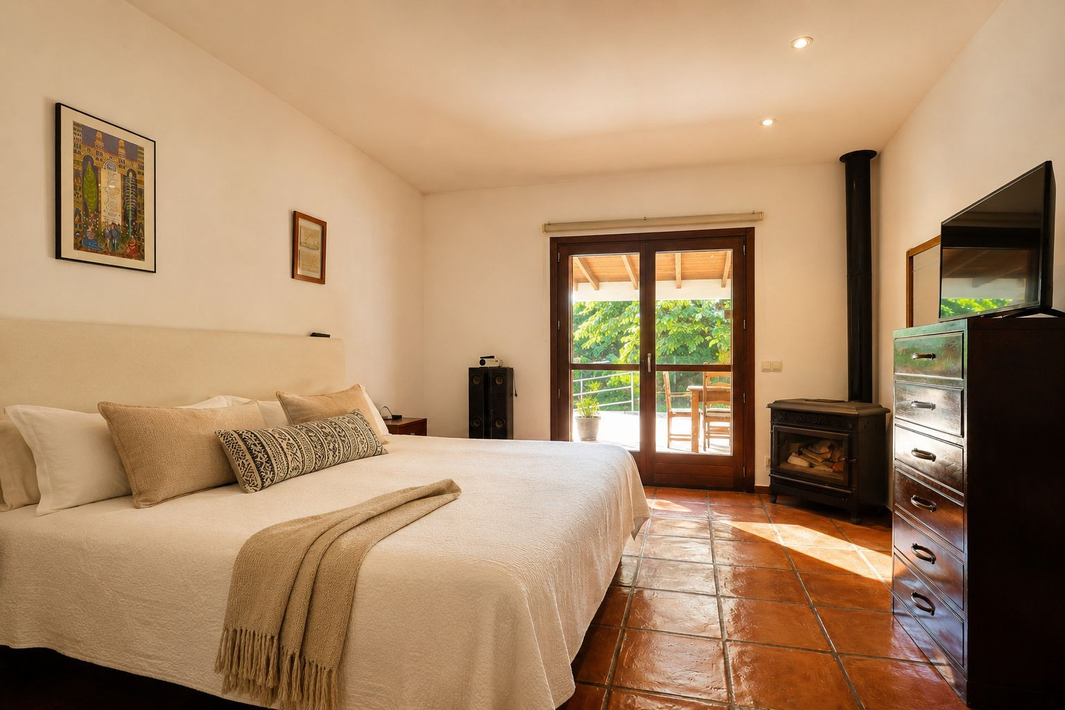 Chambre lumineuse avec un grand lit, des carreaux de terre cuite traditionnels et un poêle à bois. Une double porte vitrée s'ouvre sur une terrasse extérieure verdoyante. Chambre lumineuse avec un grand lit, des carreaux de terre cuite traditionnels et un poêle à bois. Une double porte vitrée s'ouvre sur une terrasse extérieure verdoyante.