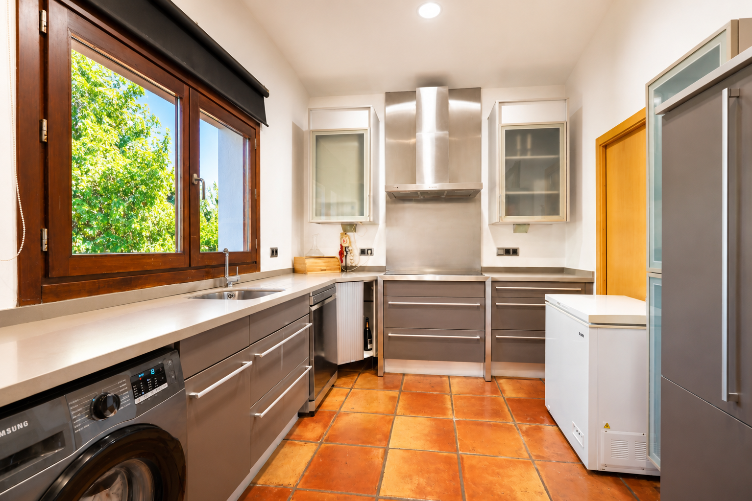 Cuisine fonctionnelle avec lave-linge intégré, sol en terre cuite artisanale et grande fenêtre en bois offrant une vue sur le jardin. Cuisine fonctionnelle avec lave-linge intégré, sol en terre cuite artisanale et grande fenêtre en bois offrant une vue sur le jardin.