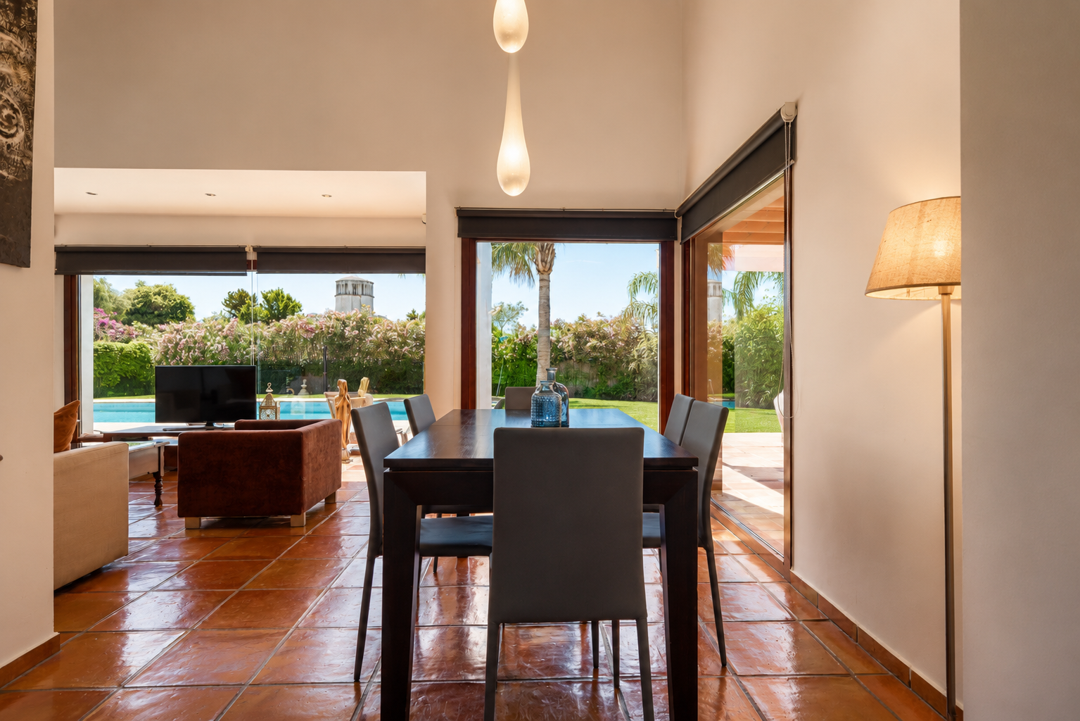 Espace salle à manger avec table en bois foncé et carrelage en terre cuite. De larges baies vitrées inondent la pièce de lumière naturelle et offrent une vue sur le jardin, la piscine et une tour. Espace salle à manger avec table en bois foncé et carrelage en terre cuite. De larges baies vitrées inondent la pièce de lumière naturelle et offrent une vue sur le jardin, la piscine et une tour.