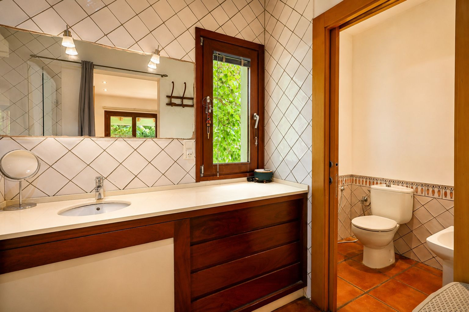 Meuble-lavabo avec vasque intégrée sur plan de travail clair et meuble en bois foncé. Le grand miroir avec appliques et la fenêtre en bois offrent une vue sur la verdure. Meuble-lavabo avec vasque intégrée sur plan de travail clair et meuble en bois foncé. Le grand miroir avec appliques et la fenêtre en bois offrent une vue sur la verdure.