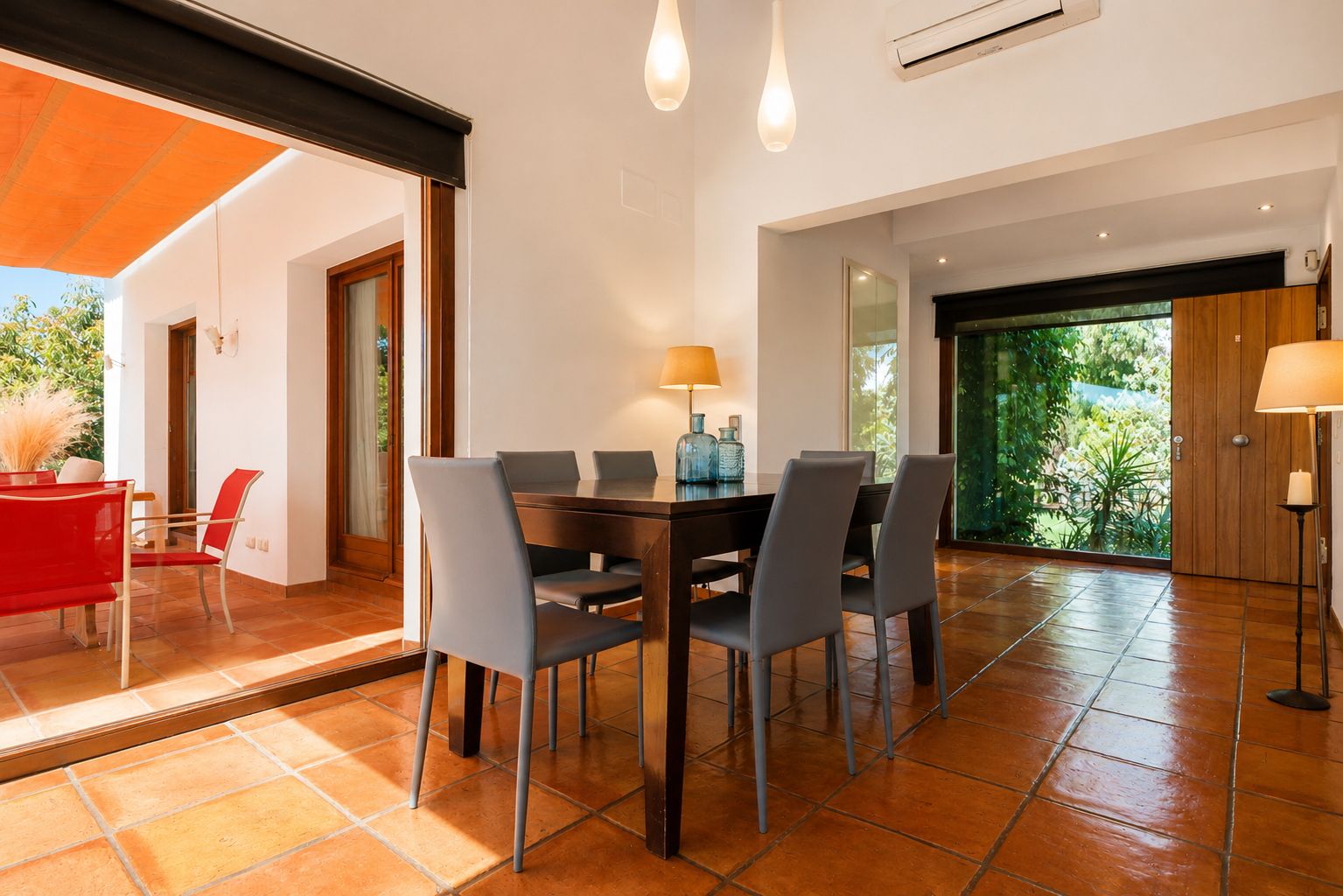 Salle à manger lumineuse avec table en bois foncé et six chaises grises, sur un sol en terre cuite. De larges baies vitrées s'ouvrent sur une terrasse extérieure ombragée et un jardin verdoyant. Salle à manger lumineuse avec table en bois foncé et six chaises grises, sur un sol en terre cuite. De larges baies vitrées s'ouvrent sur une terrasse extérieure ombragée et un jardin verdoyant.
