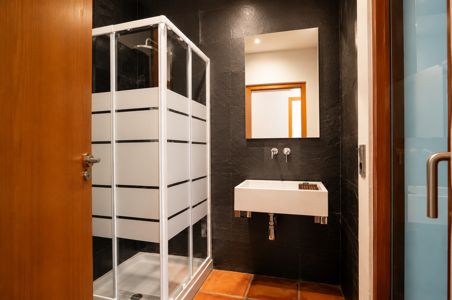 Salle de bain avec cabine de douche à bandes dépolies, lavabo rectangulaire suspendu et carrelage mural texturé foncé. Le sol est en carreaux de terre cuite. Salle de bain avec cabine de douche à bandes dépolies, lavabo rectangulaire suspendu et carrelage mural texturé foncé. Le sol est en carreaux de terre cuite.