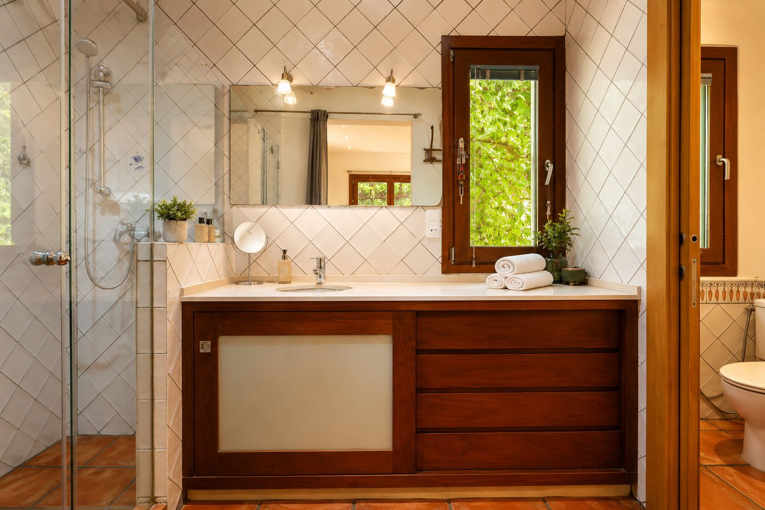 Salle de bain avec douche vitrée, meuble-lavabo en bois foncé et plan de travail blanc. Fenêtre donnant sur la verdure, carreaux blancs en losange et sols en terre cuite artisanale. Salle de bain avec douche vitrée, meuble-lavabo en bois foncé et plan de travail blanc. Fenêtre donnant sur la verdure, carreaux blancs en losange et sols en terre cuite artisanale.