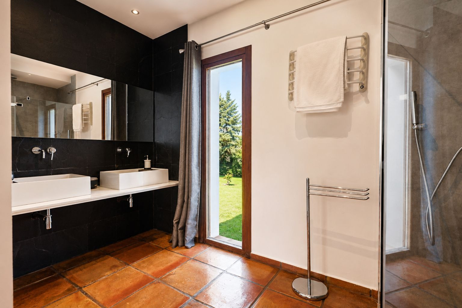 Salle de bain spacieuse avec carrelage en terre cuite, double vasque rectangulaire blanche sur un mur carrelé noir. Grande fenêtre offrant une vue sur le jardin et douche vitrée. Salle de bain spacieuse avec carrelage en terre cuite, double vasque rectangulaire blanche sur un mur carrelé noir. Grande fenêtre offrant une vue sur le jardin et douche vitrée.