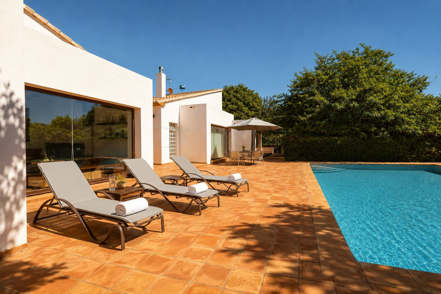 Vaste terrasse carrelée en terre cuite avec piscine, trois chaises longues et un espace repas ombragé. La façade blanche est agrémentée de grandes fenêtres reflétant la végétation luxuriante. Vaste terrasse carrelée en terre cuite avec piscine, trois chaises longues et un espace repas ombragé. La façade blanche est agrémentée de grandes fenêtres reflétant la végétation luxuriante.