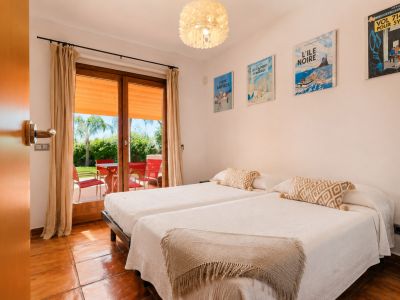 Chambre double avec carrelage en terre cuite et accès direct à une terrasse extérieure. La porte vitrée mène à un jardin verdoyant avec mobilier rouge et palmier.