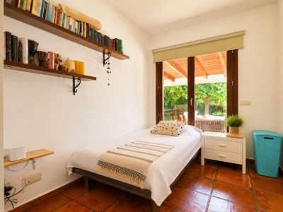 Chambre individuelle avec carrelage en terre cuite, murs blancs et accès direct à une terrasse couverte par une grande porte vitrée en bois. Des étagères murales avec des livres sont visibles.