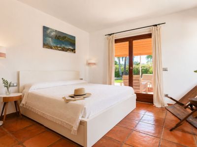 Chambre lumineuse avec lit blanc, sols en terre cuite artisanale et accès direct à une terrasse extérieure offrant vue sur le jardin.