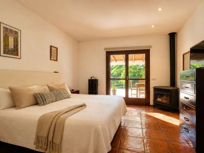 Chambre lumineuse avec un grand lit, des carreaux de terre cuite traditionnels et un poêle à bois. Une double porte vitrée s'ouvre sur une terrasse extérieure verdoyante.