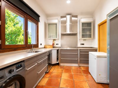 Cuisine fonctionnelle avec lave-linge intégré, sol en terre cuite artisanale et grande fenêtre en bois offrant une vue sur le jardin.