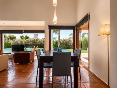 Espace salle à manger avec table en bois foncé et carrelage en terre cuite. De larges baies vitrées inondent la pièce de lumière naturelle et offrent une vue sur le jardin, la piscine et une tour.