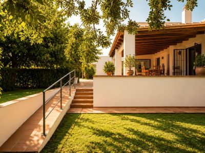 Naya couverte avec poutres en bois apparentes et coin repas extérieur, donnant sur une pelouse entretenue et un jardin méditerranéen, accessible par rampe et escaliers.