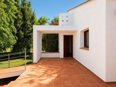 Patio d'entrée principal couvert avec carrelage en terre cuite et éclairage encastré. Une rampe accessible mène au jardin verdoyant, s'harmonisant avec la façade blanche.