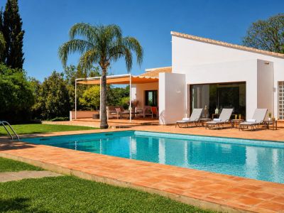 Piscine carrelée de bleu et entourée de terre cuite, bordée d'une pelouse verte. Des chaises longues sont disposées devant la façade blanche de la maison, près d'une pergola ombragée.