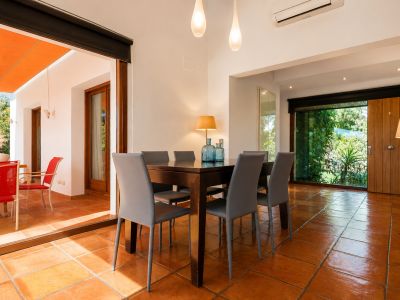 Salle à manger lumineuse avec table en bois foncé et six chaises grises, sur un sol en terre cuite. De larges baies vitrées s'ouvrent sur une terrasse extérieure ombragée et un jardin verdoyant.