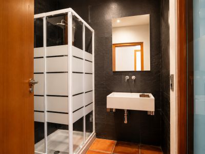 Salle de bain avec cabine de douche à bandes dépolies, lavabo rectangulaire suspendu et carrelage mural texturé foncé. Le sol est en carreaux de terre cuite.