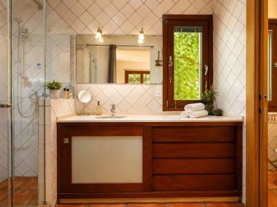 Salle de bain avec douche vitrée, meuble-lavabo en bois foncé et plan de travail blanc. Fenêtre donnant sur la verdure, carreaux blancs en losange et sols en terre cuite artisanale.